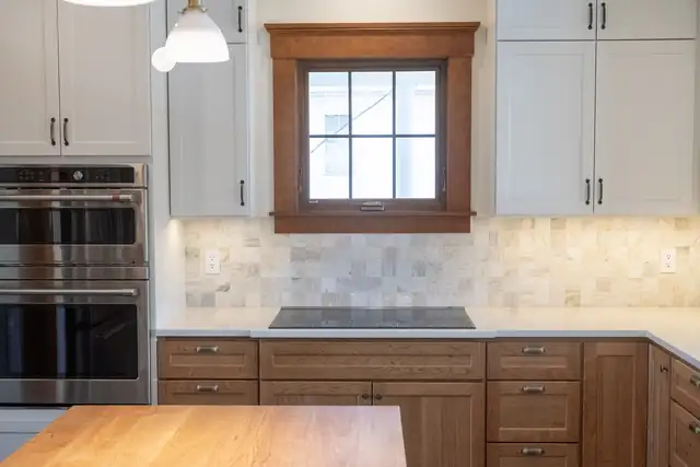 Kitchen with warm wood lower cabinets, white uppers, double wall ovens and stone tile backsplash under a wood-trimmed window